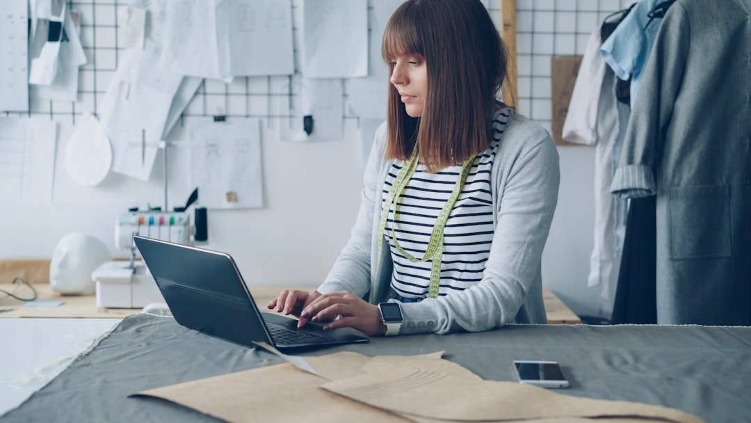 A seamstress works on her laptop in a bright workspace.